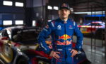 Race car driver in a blue Red Bull suit standing with hands on hips in a dim garage near a colorful race car.