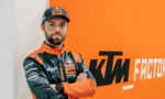 Man in orange and dark blue KTM Red Bull racing jacket stands with arms crossed in front of an orange wall with 'KTM Factory Racing' logo.