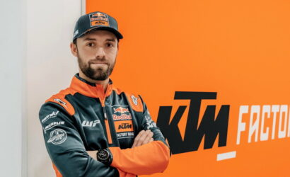 Man in orange and dark blue KTM Red Bull racing jacket stands with arms crossed in front of an orange wall with 'KTM Factory Racing' logo.