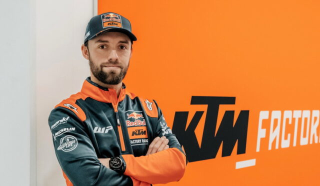 Man in orange and dark blue KTM Red Bull racing jacket stands with arms crossed in front of an orange wall with 'KTM Factory Racing' logo.