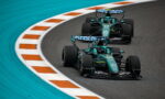 Two green Formula 1 cars race through a sharp right-hand corner, hugging the orange and white curb on a teal runoff area behind them