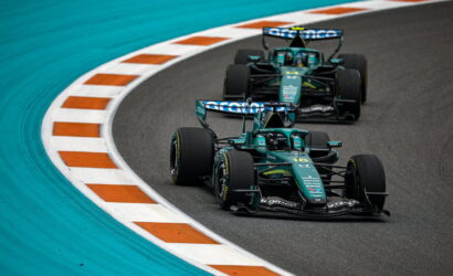 Two green Formula 1 cars race through a sharp right-hand corner, hugging the orange and white curb on a teal runoff area behind them