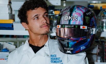Male researcher in a white lab coat gazes at a colorful, logo-covered racing helmet in a lab setting.