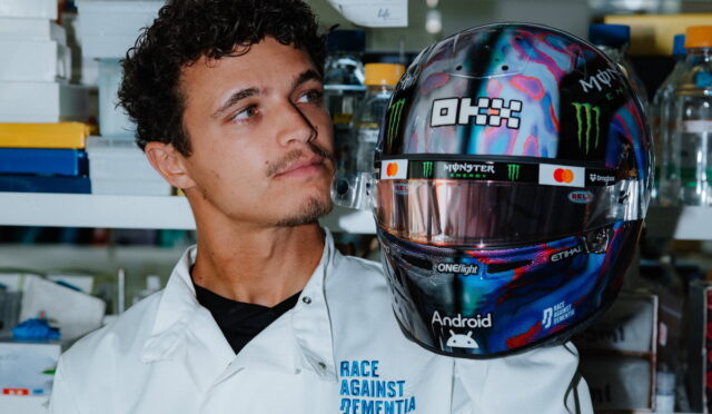 Male researcher in a white lab coat gazes at a colorful, logo-covered racing helmet in a lab setting.