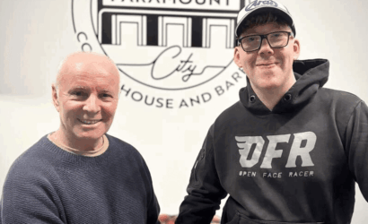 Two men smiling indoors in front of a circular logo on the wall; an older man in a blue sweater and a younger man in a black hoodie and cap stand side by side.