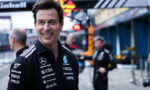 Smiling man in a black sponsor-logo polo stands in a pit lane with crew members blurred in the background.