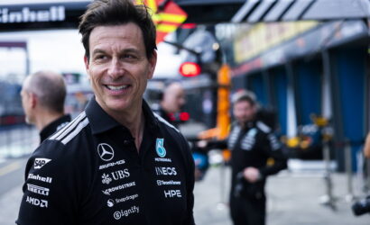 Smiling man in a black sponsor-logo polo stands in a pit lane with crew members blurred in the background.