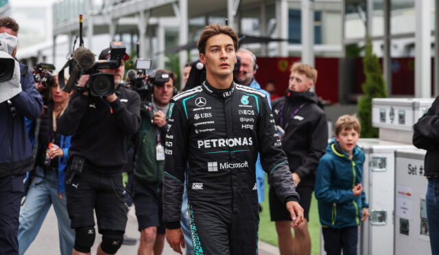 Formula 1 driver in a Petronas Mercedes suit walks past a crowd of photographers at a race event.