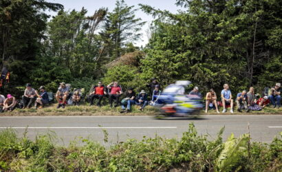 Group of spectators sitting along a grassy verge beside a road, watching a blurred racing motorcycle pass by in the foreground, with trees in the background.