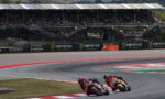Two riders on sport motorcycles lean into a left-hand turn on a race track, with grandstands and Monster Energy banners in the background.
