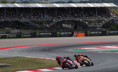 Two riders on sport motorcycles lean into a left-hand turn on a race track, with grandstands and Monster Energy banners in the background.