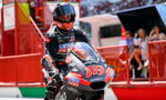 Motorcycle racer in a black suit and orange helmet on a racing bike number 78, at a track with red barriers and spectators in the background.