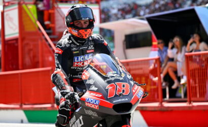 Motorcycle racer in a black suit and orange helmet on a racing bike number 78, at a track with red barriers and spectators in the background.