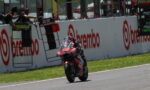 Motorcycle racer in red and black racing suit rides a bike at speed along a track, leaning into a turn with sponsor banners on the wall behind him.