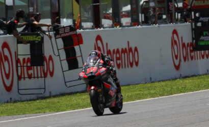 Motorcycle racer in red and black racing suit rides a bike at speed along a track, leaning into a turn with sponsor banners on the wall behind him.