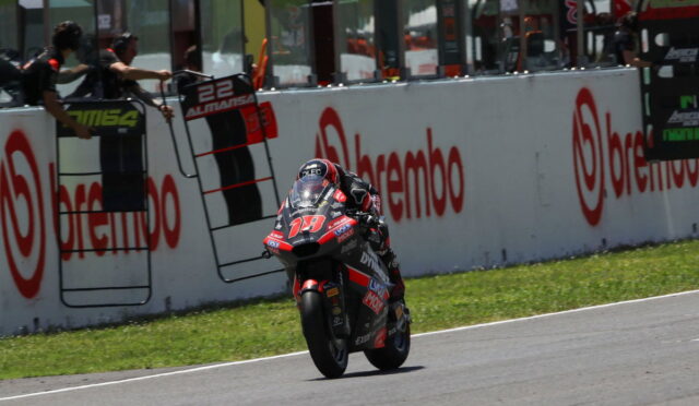 Motorcycle racer in red and black racing suit rides a bike at speed along a track, leaning into a turn with sponsor banners on the wall behind him.
