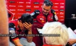 Two racing team members in red uniforms in a pit area; one man covers his face with his hand while the other stands nearby with a clipboard, sponsor banners in the background. Caption reads: 'There's a screw that's giving me nerve problems.'