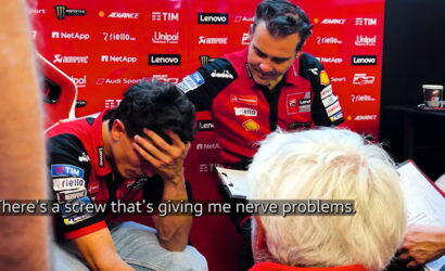 Two racing team members in red uniforms in a pit area; one man covers his face with his hand while the other stands nearby with a clipboard, sponsor banners in the background. Caption reads: 'There's a screw that's giving me nerve problems.'