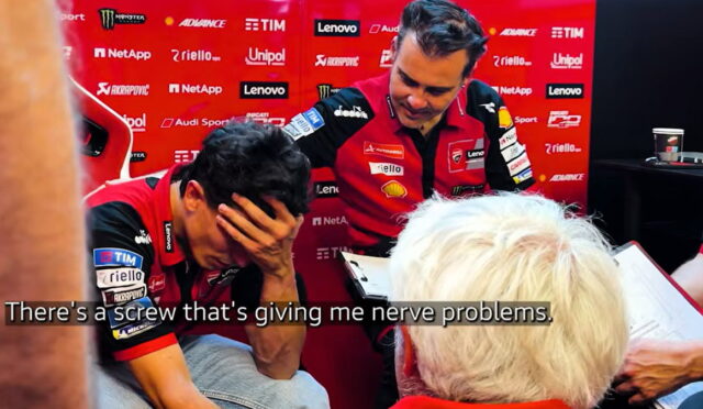 Two racing team members in red uniforms in a pit area; one man covers his face with his hand while the other stands nearby with a clipboard, sponsor banners in the background. Caption reads: 'There's a screw that's giving me nerve problems.'