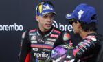 Two motorcycle racers in full racing suits converse, one facing the camera wearing a blue Michelin cap and sunglasses on his head.
