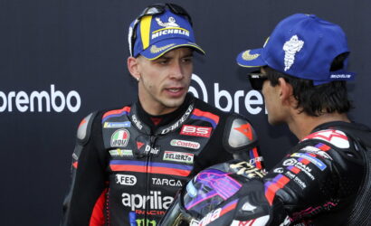 Two motorcycle racers in full racing suits converse, one facing the camera wearing a blue Michelin cap and sunglasses on his head.