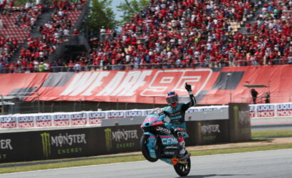 Moto racer performs a wheelie on a teal racing bike as spectators cheer in the stands behind him.