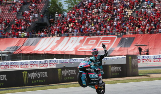 Moto racer performs a wheelie on a teal racing bike as spectators cheer in the stands behind him.