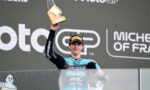 MotoGP rider on podium raising a wooden trophy above his head during a race celebration, with sponsor logos in the background