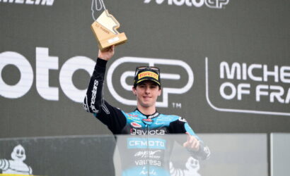 MotoGP rider on podium raising a wooden trophy above his head during a race celebration, with sponsor logos in the background