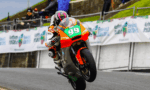 Rider on a bright orange-green race bike #99 performs a wheelie on a track with spectators and banners in the background.