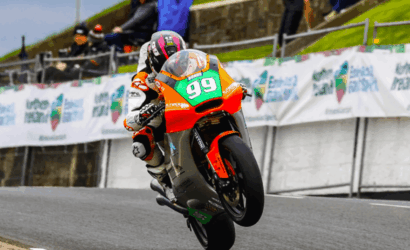 Rider on a bright orange-green race bike #99 performs a wheelie on a track with spectators and banners in the background.