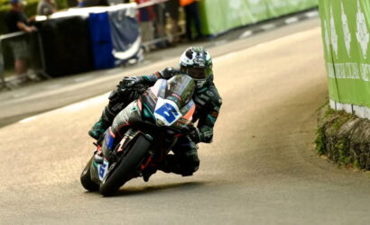 Motorbike racer in teal-green leans into a tight turn on a street circuit, number 3 on the bike, helmeted in white.