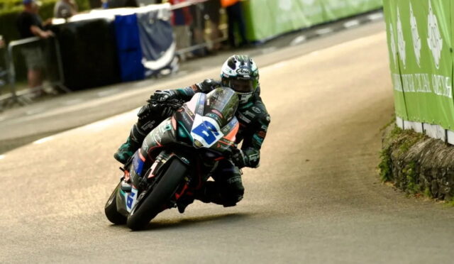 Motorbike racer in teal-green leans into a tight turn on a street circuit, number 3 on the bike, helmeted in white.
