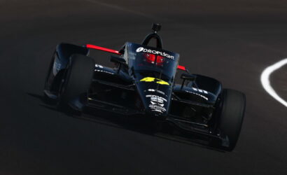 Front-left view of a dark blue formula-style race car on a race track, with sponsor decals and a red roll hoop behind the cockpit.