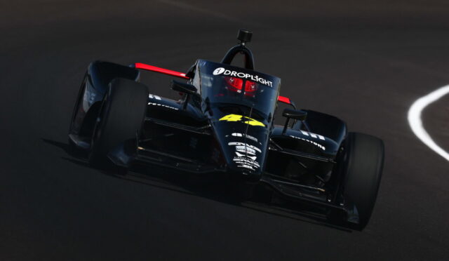 Front-left view of a dark blue formula-style race car on a race track, with sponsor decals and a red roll hoop behind the cockpit.