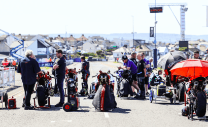 Racetrack pit area with multiple racing motorcycles and crew members preparing under sunny skies near a coastal town.