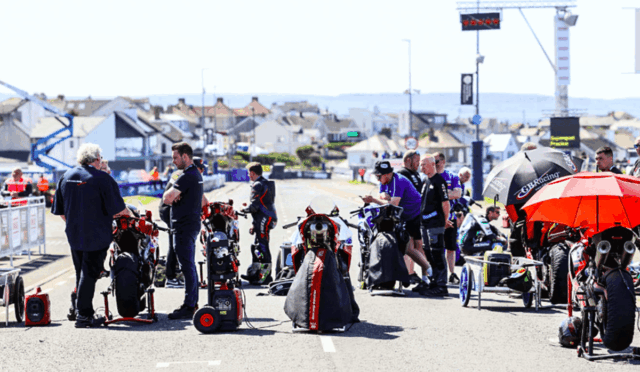 Racetrack pit area with multiple racing motorcycles and crew members preparing under sunny skies near a coastal town.
