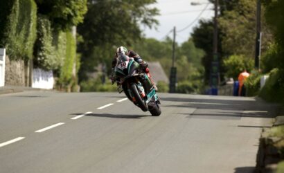 Motorcycle racer leaning sharply into a turn on a quiet rural road, wearing a black helmet and teal-red suit, number 10 on the bike.