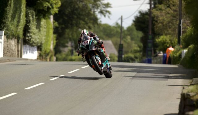 Motorcycle racer leaning sharply into a turn on a quiet rural road, wearing a black helmet and teal-red suit, number 10 on the bike.
