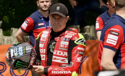 Race car or motorcycle rider in red Honda suit holds helmet, standing outdoors with crew members nearby.