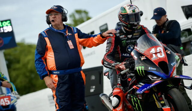 Moto racer in full suit and helmet on a race bike, while a crew member in blue‑orange jumpsuit steadies him from the pit area with a headset on.