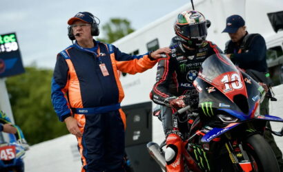 Race bike and rider in full leathers and helmet at the pit, trainer in blue‑orange suit offers support beside the bike