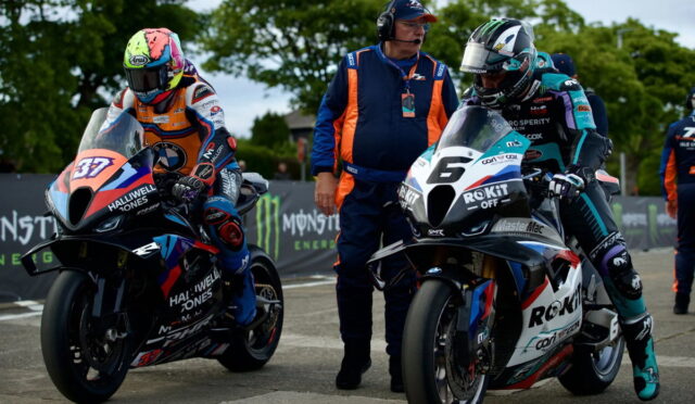 Two motorcycle racers in colorful leathers line up on a track, with a marshal in an orange-and-navy suit standing between them.