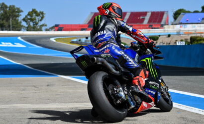 Moto racer on a blue Yamaha bike leaning into a turn on a race track; Monster Energy livery and red helmet visible.