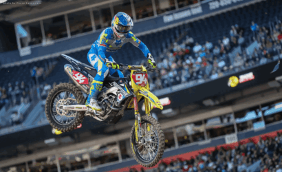 Motocross rider in blue gear mid-air on a yellow dirt bike during an indoor stadium race with spectators watching.