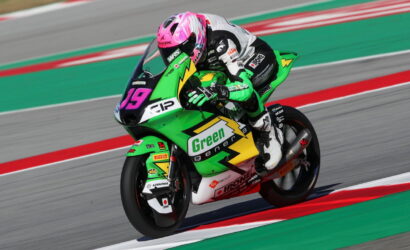 MotoGP rider in a pink helmet leans into a corner on a bright green race bike with 'Green energy' branding, number 19 visible on the front plate.