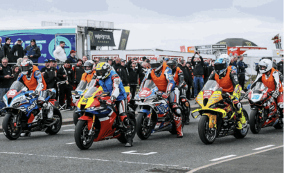 Group of motorcycle racers lined up on a track, colorful leathers and bikes ready for a start, with spectators in the background.