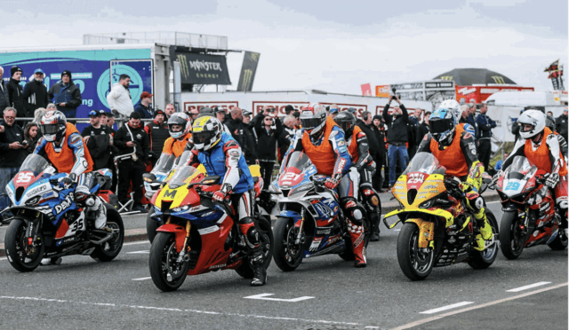 Group of motorcycle racers lined up on a track, colorful leathers and bikes ready for a start, with spectators in the background.