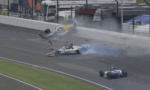 Race car crashes on track, flipping into the air with debris and smoke around the scene at a racing circuit behind a barrier.