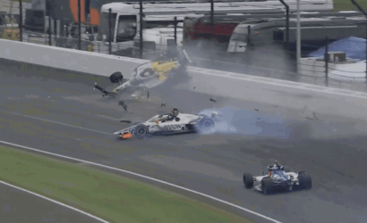 Race car crashes on track, flipping into the air with debris and smoke around the scene at a racing circuit behind a barrier.